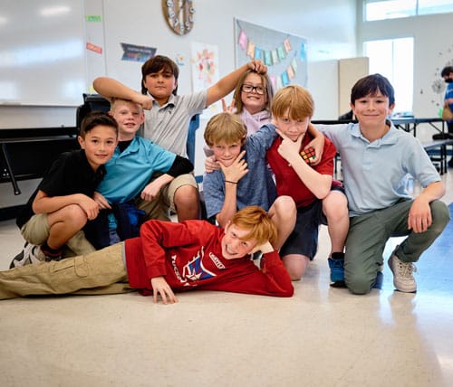 A group of smiling elementary students posing playfully on the classroom floor, with desks and colorful decorations in the background.
